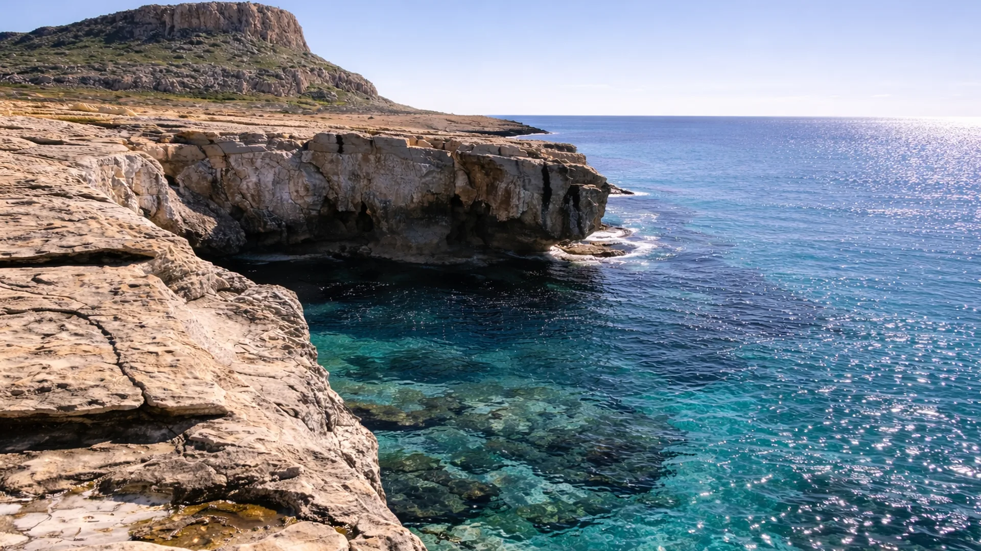 Rugged limestone cliffs at Cape Greco overlooking clear turquoise Mediterranean waters under a bright blue sky in Cyprus.