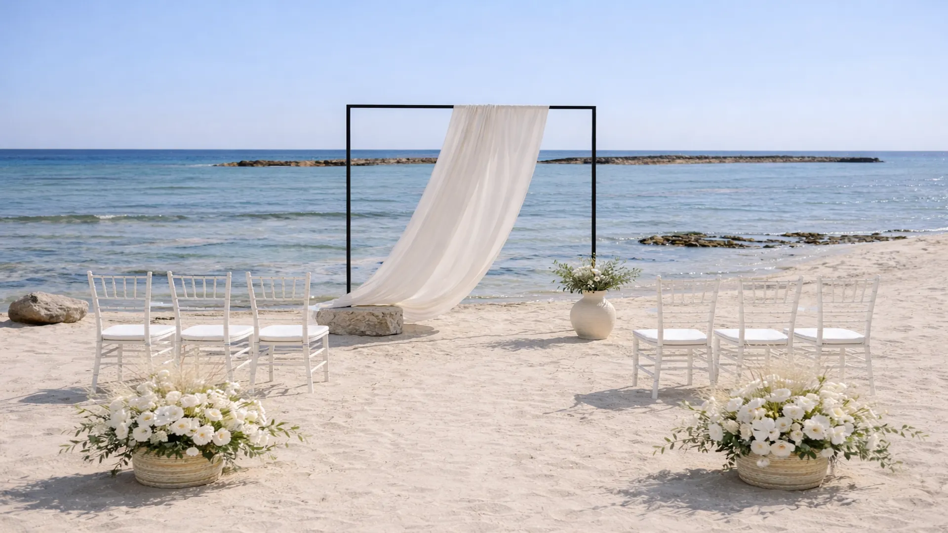 Minimalist beach wedding ceremony in Cyprus featuring a black metal arch with soft white draping, natural florals, and an open Mediterranean Sea backdrop.