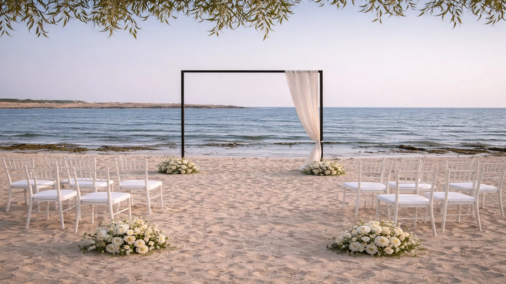 Minimalist beach wedding ceremony in Cyprus with a black frame arch, white chairs, and soft floral accents overlooking the Mediterranean Sea at sunset.